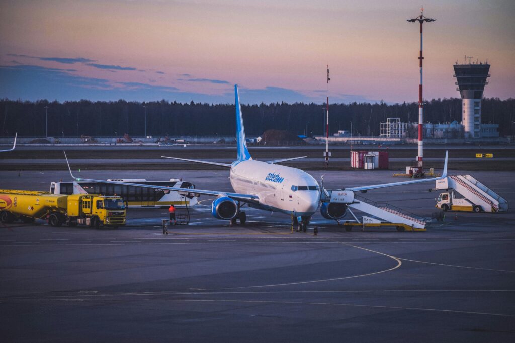 A commercial airplane parked at an airport gate during twilight, with a fuel truck refueling it, a passenger boarding staircase positioned nearby, and an air traffic control tower in the background