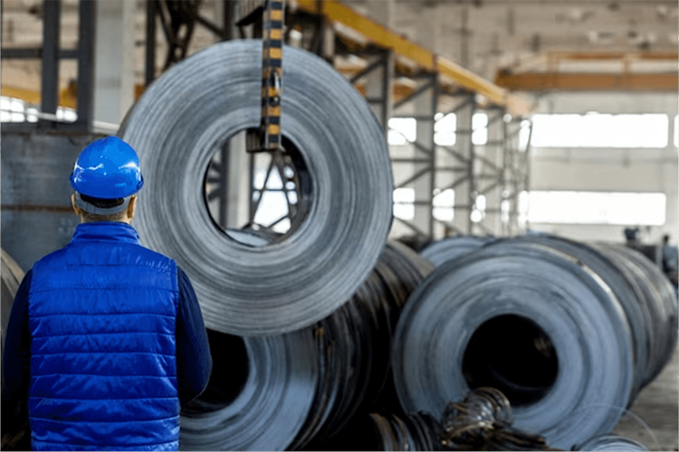 Industrial worker in a blue safety vest and helmet observing large steel coils being moved by a crane in a factory setting