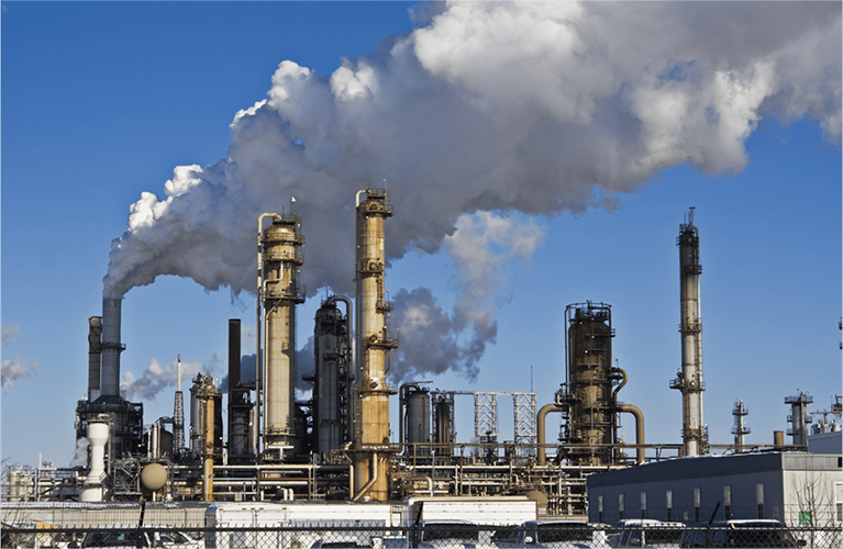 Large industrial oil refinery with multiple chimneys emitting thick white smoke against a clear blue sky