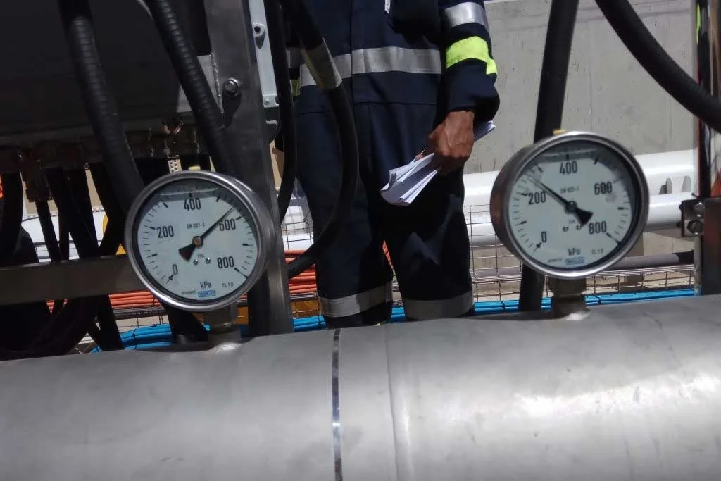 Close-up of two pressure gauges on industrial equipment, with a technician in protective uniform holding documents in the background