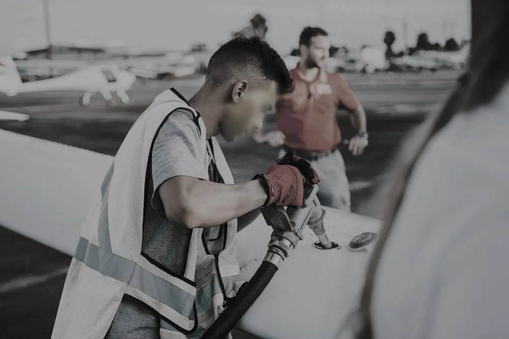Worker wearing safety vest refueling an aircraft on the tarmac, with planes and personnel in the background