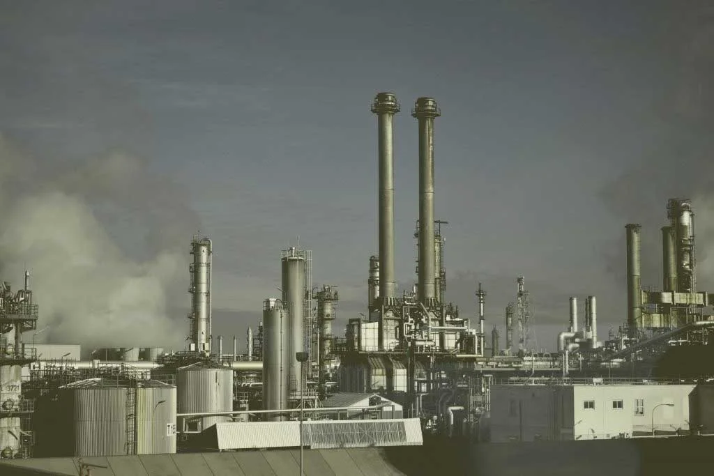 Wide view of an industrial refinery complex with multiple distillation towers, storage tanks, and smoke rising in the background
